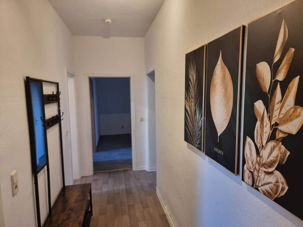 Ein Flur mit Holzboden, einem schwarzen Metallregal und gerahmten Pflanzenabbildungen an der Wand.