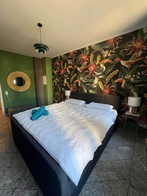 A bed with a white bedspread and blue towels, a round mirror on the wall, and a tropical-themed wallpaper. - Leipzig Alt Lindenau Apartment – Sleeps 5, Parking (Leipzig)