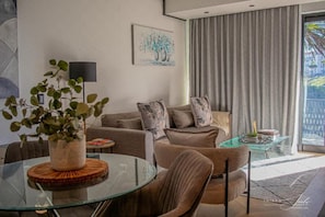 A modern living room with a glass coffee table, a sofa set, a potted plant, and a painting on the wall. - KML Nine Palms Luxury 101 (Cape Town)