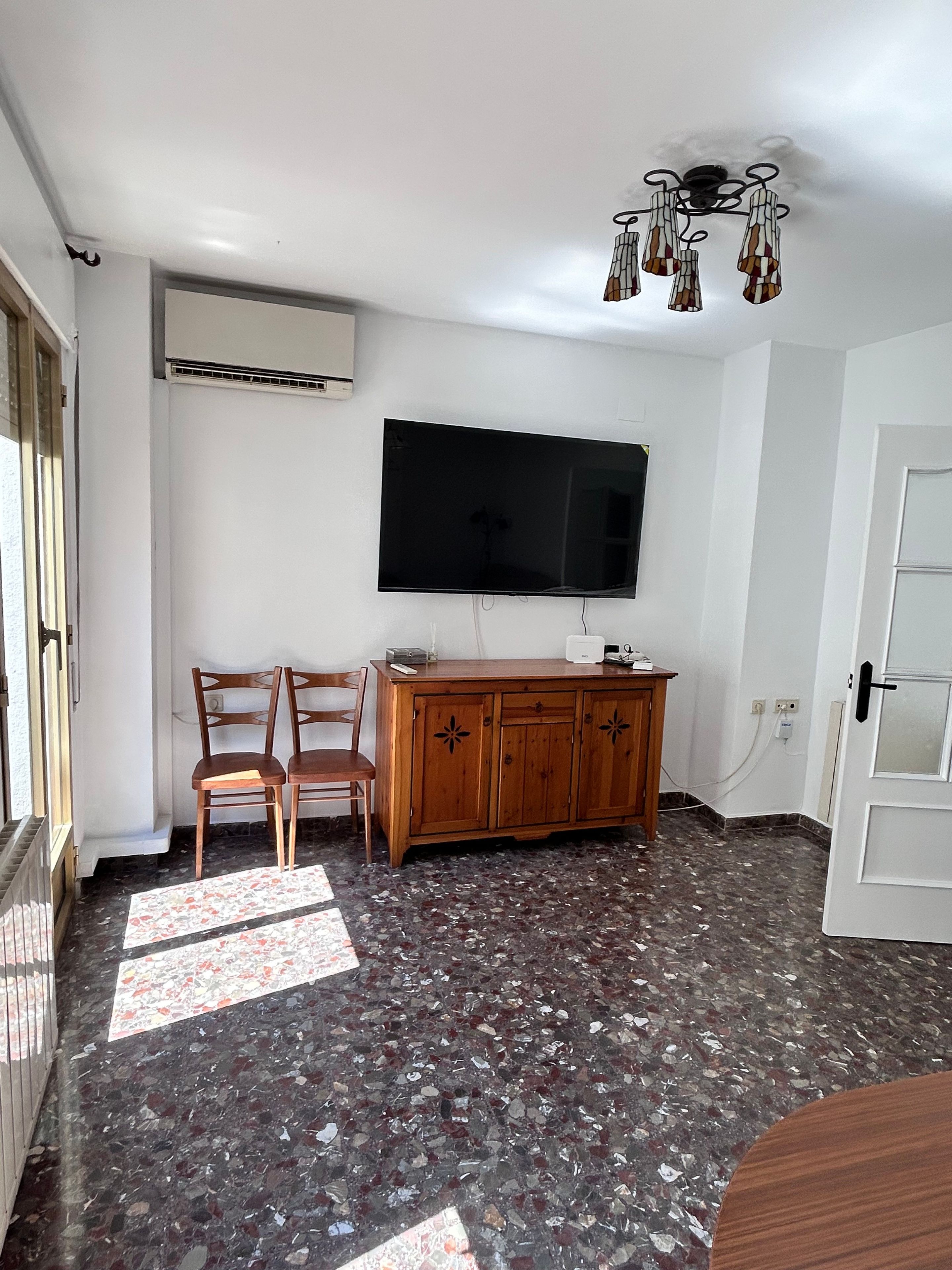 Una habitación con un televisor de pantalla plana montado en la pared, un mueble de madera, dos sillas, una alfombra estampada y suelo de terrazo.
