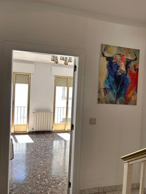 Un pasillo interior con una puerta que da a un balcón, un radiador y un cuadro de un toro colorido en la pared.