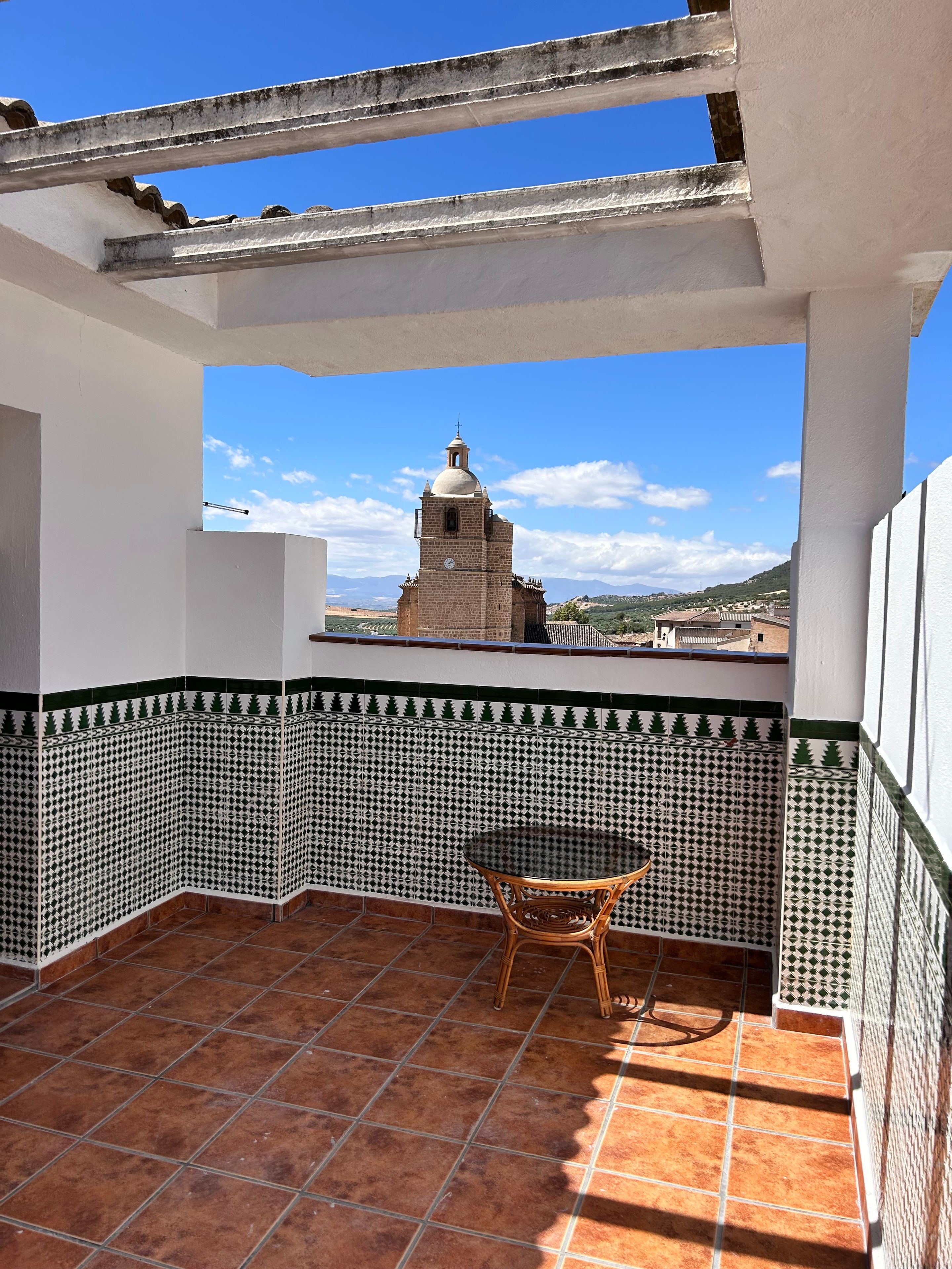 Un balcón con piso de baldosas, una mesa y vistas a una torre de iglesia.