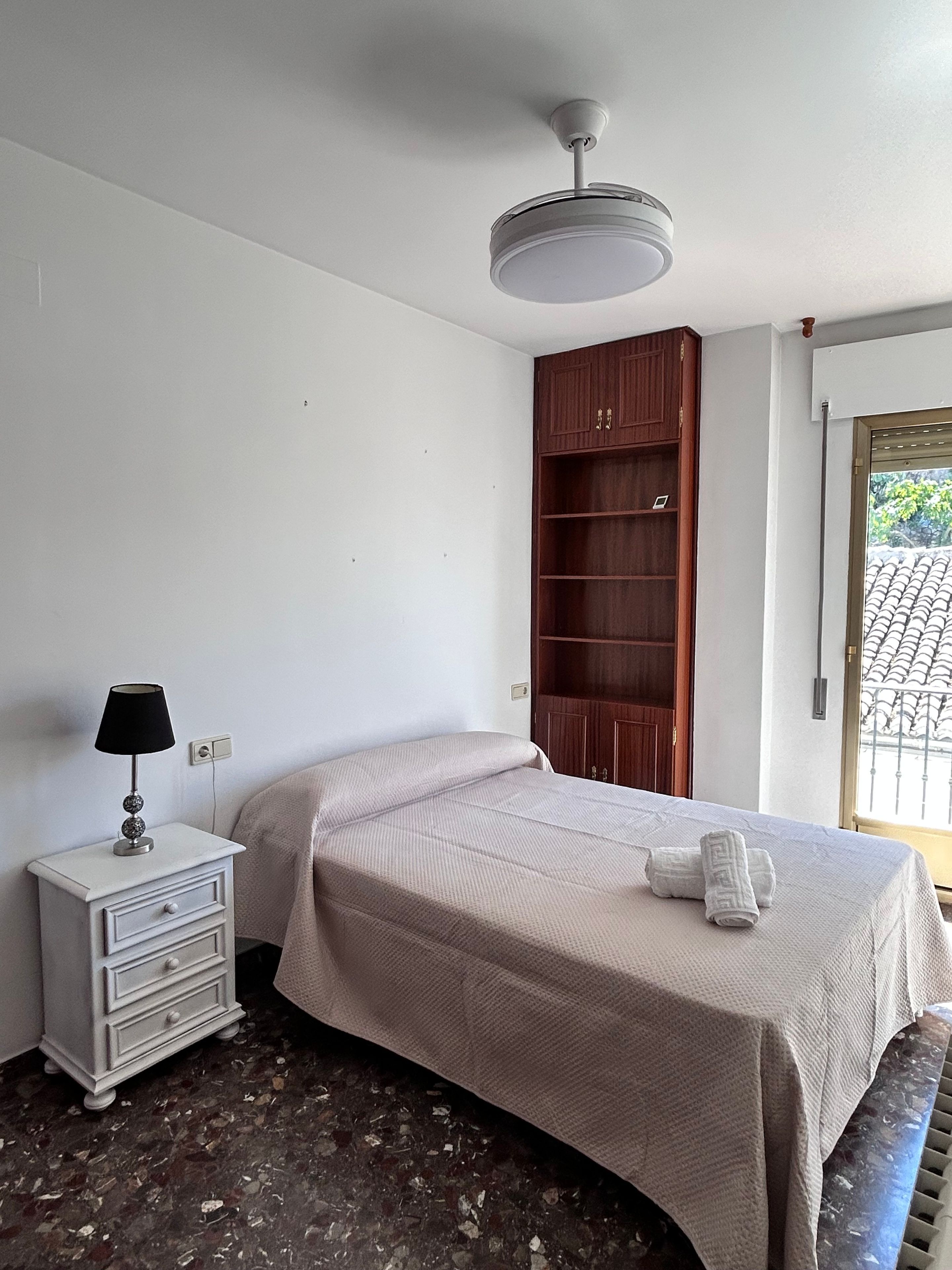 A bedroom with a bed, a nightstand, a lamp, a wardrobe, and a window with a view.