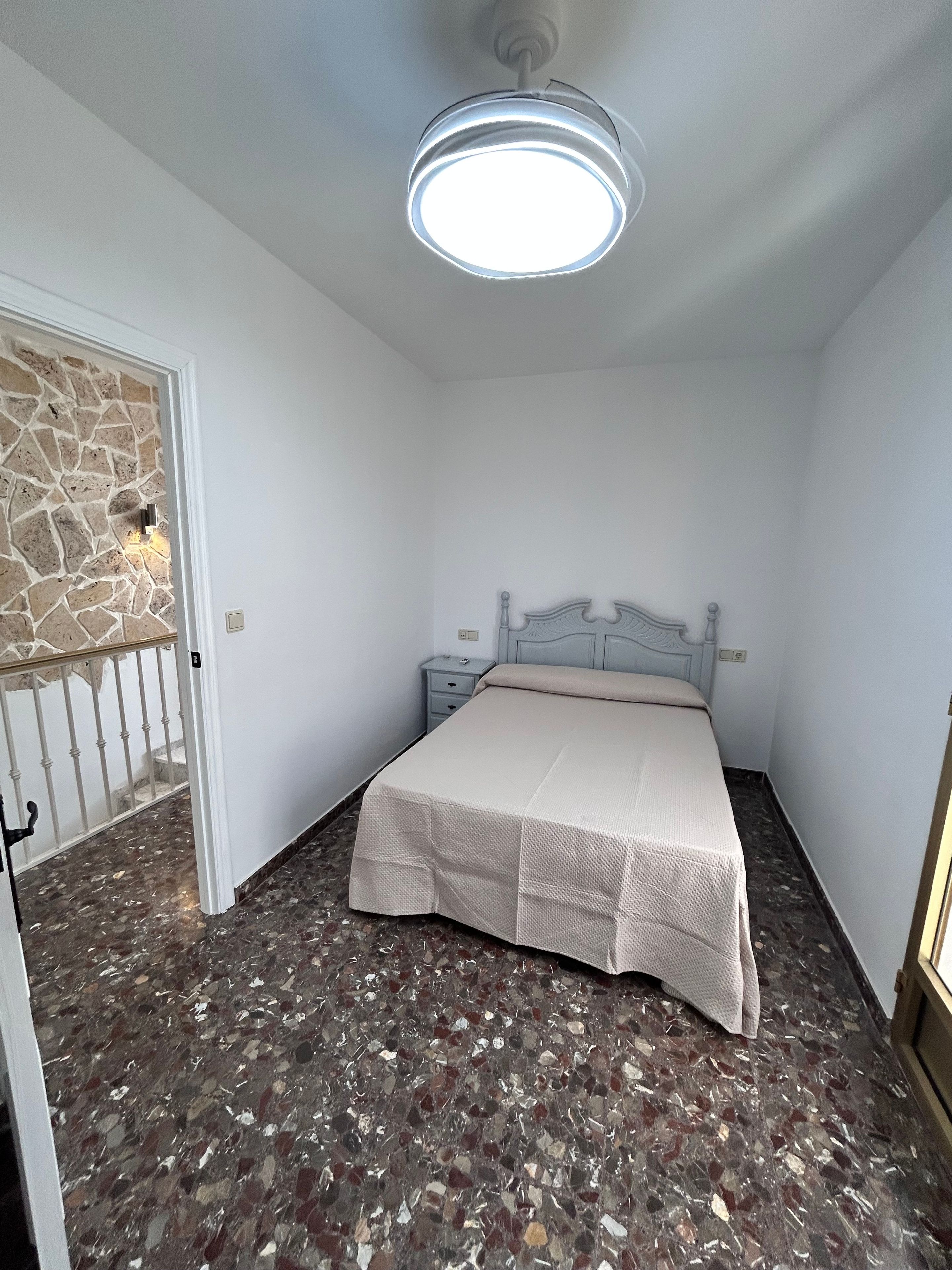 Un dormitorio con cama, mesita de noche, pared de piedra y luz de techo.