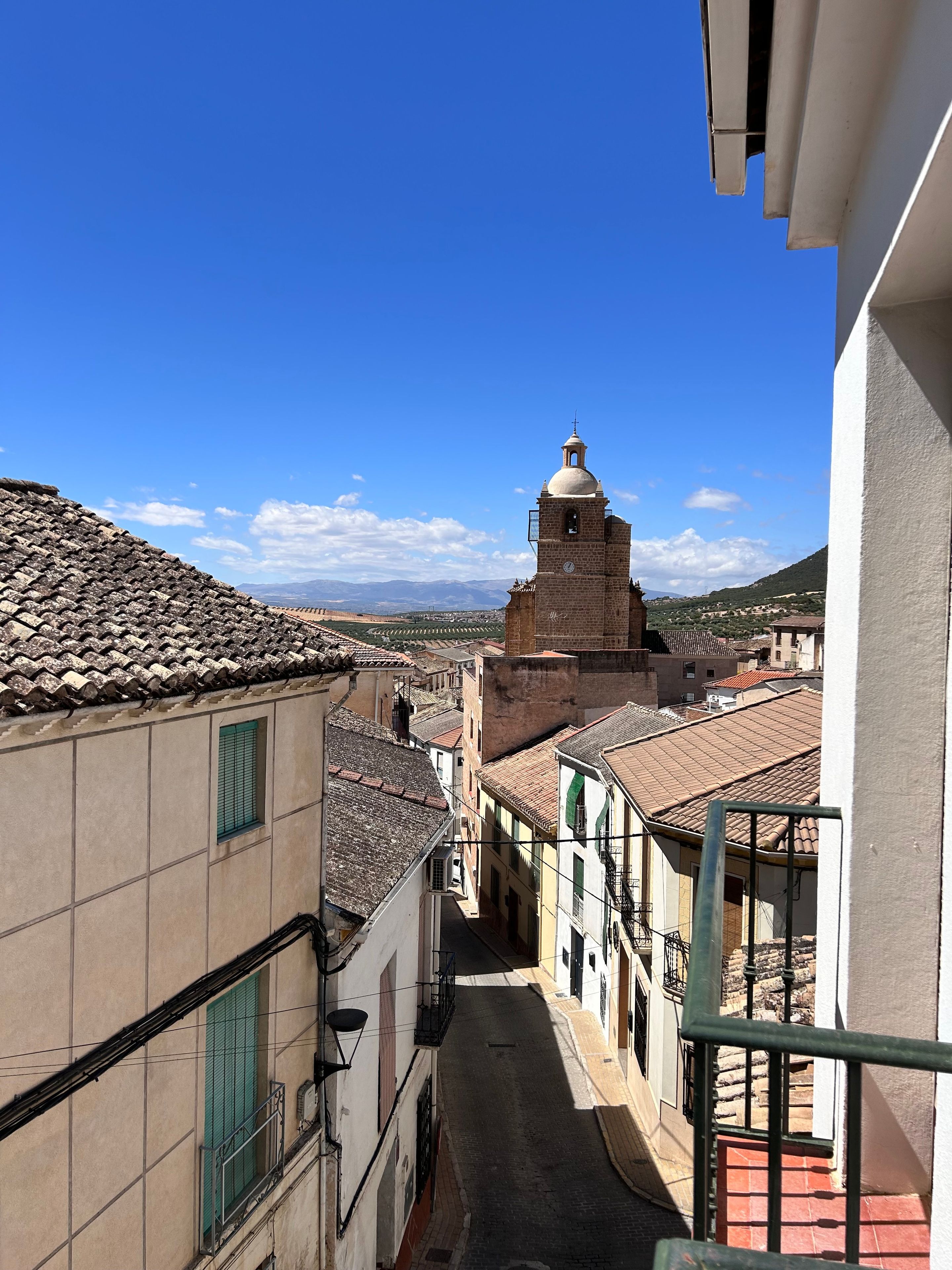 A narrow street with buildings on either side, a bell tower, and a clear blue sky.