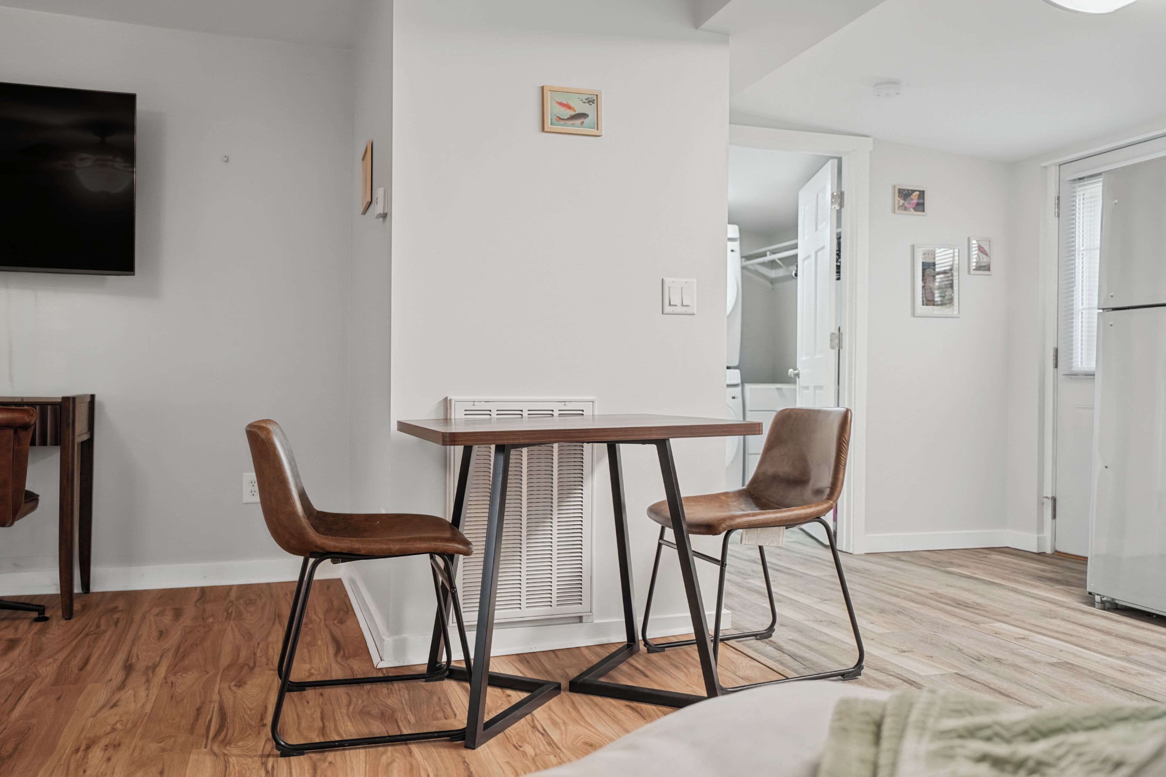Un soggiorno moderno con pavimento in legno, un tavolo da pranzo con due sedie, una TV a schermo piatto montata a parete e una piccola foto incorniciata sopra il tavolo.