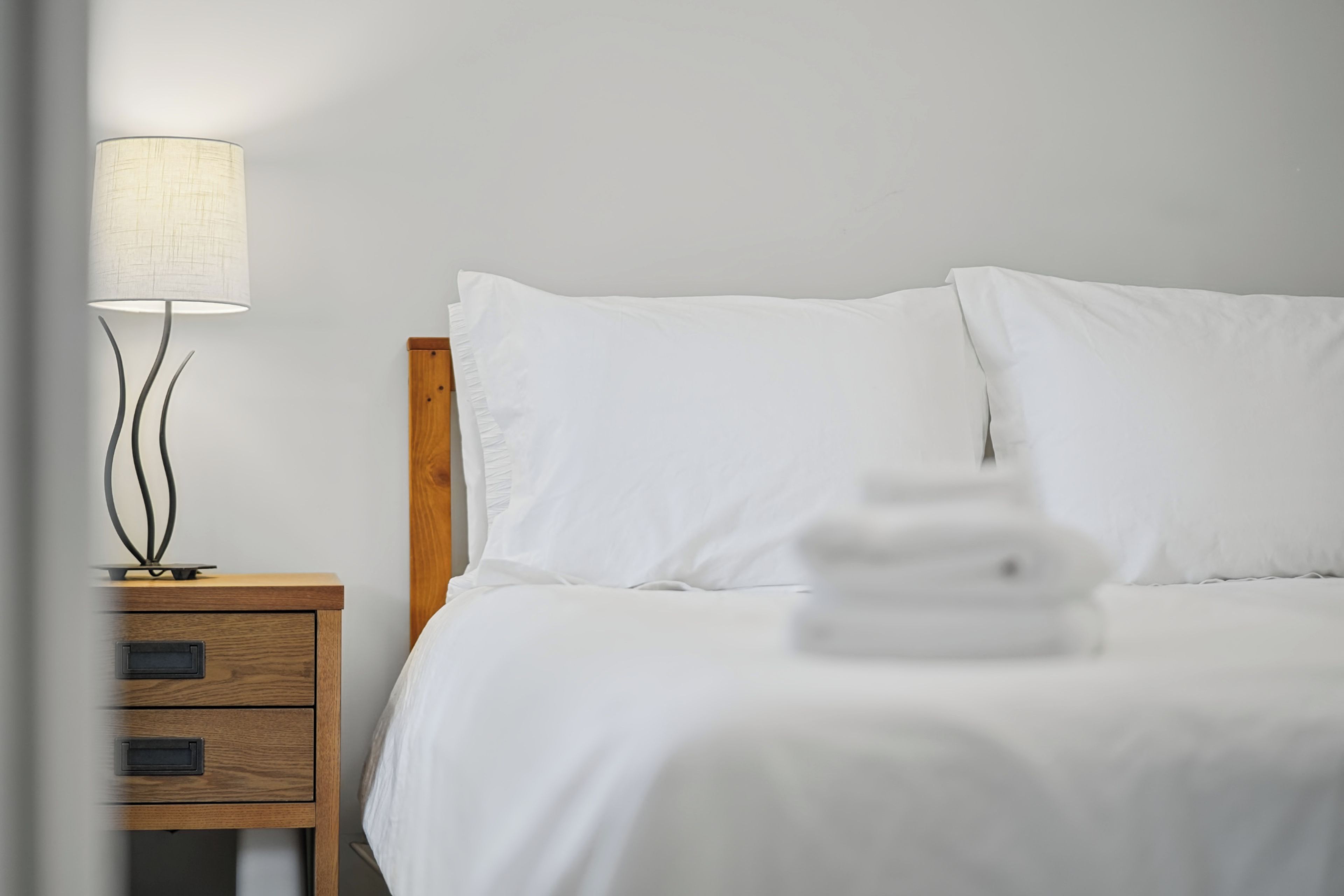 Una cama bien tendida con sábanas blancas, una mesita de noche con lámpara y un mueble cajonero de madera.