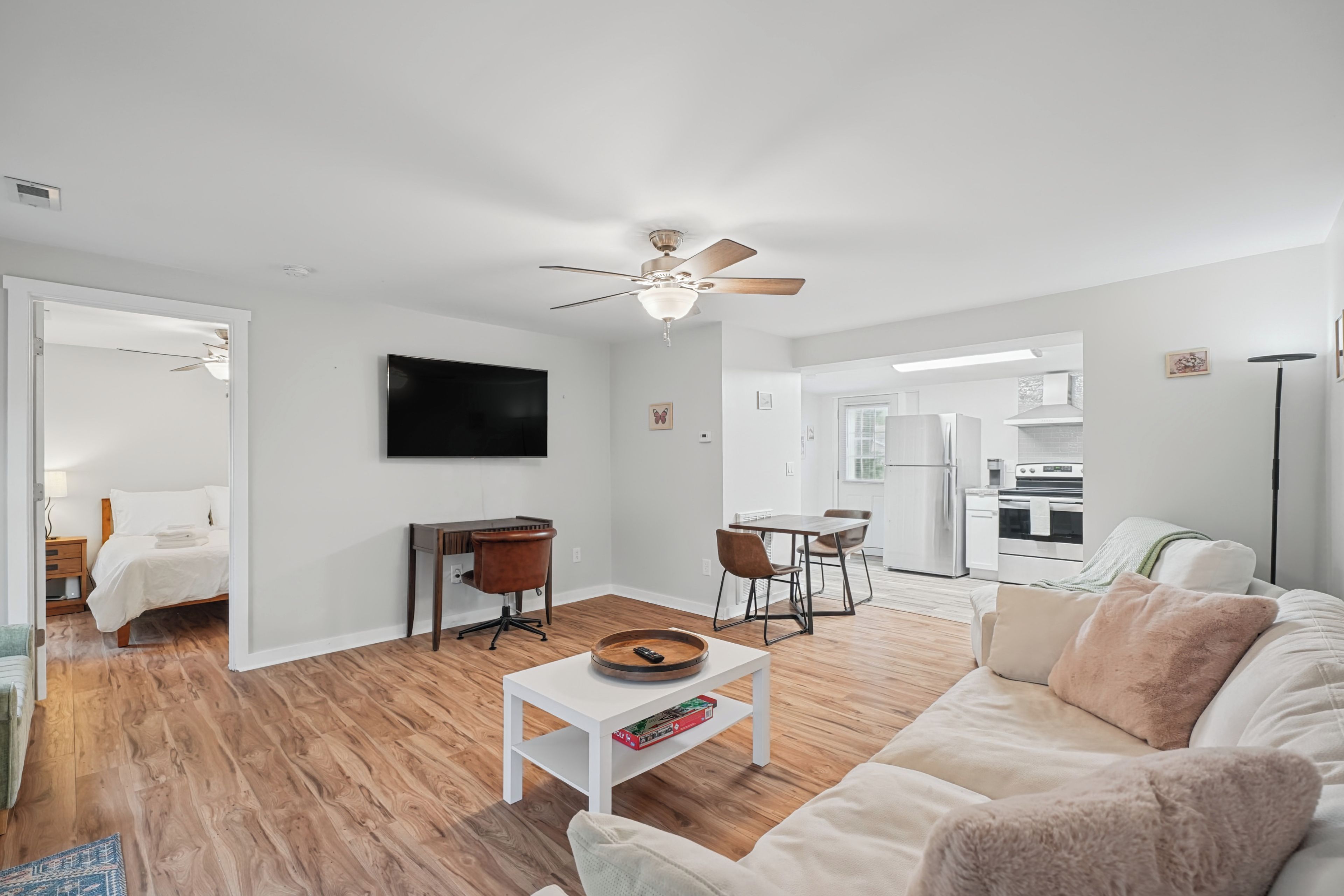 A modern living room with a flat-screen TV, a ceiling fan, a dining area with a table and chairs, and a kitchen in the background.