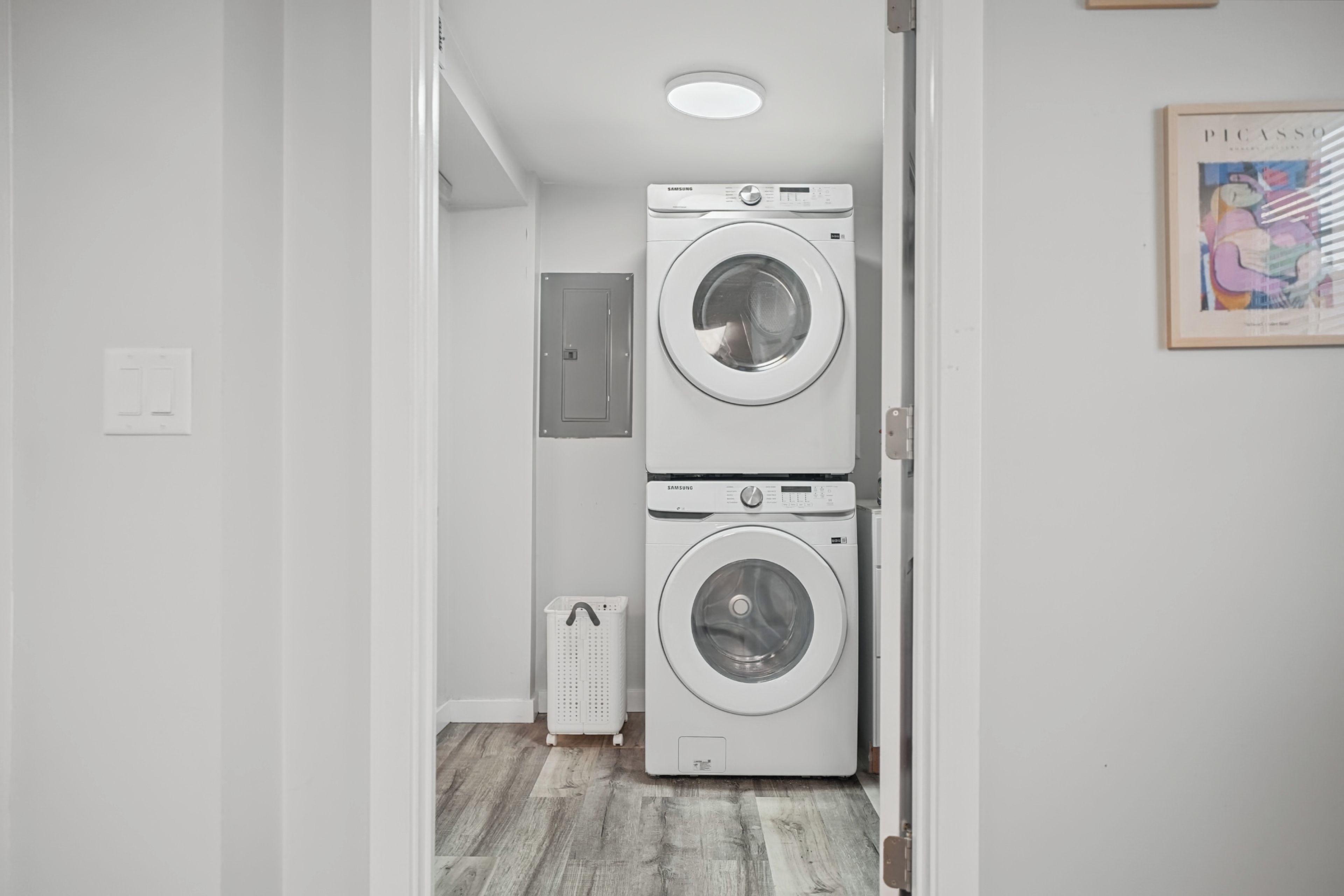 Une buanderie équipée de deux machines à laver superposées, d’un petit panier à linge blanc et d’une porte donnant sur une autre pièce.
