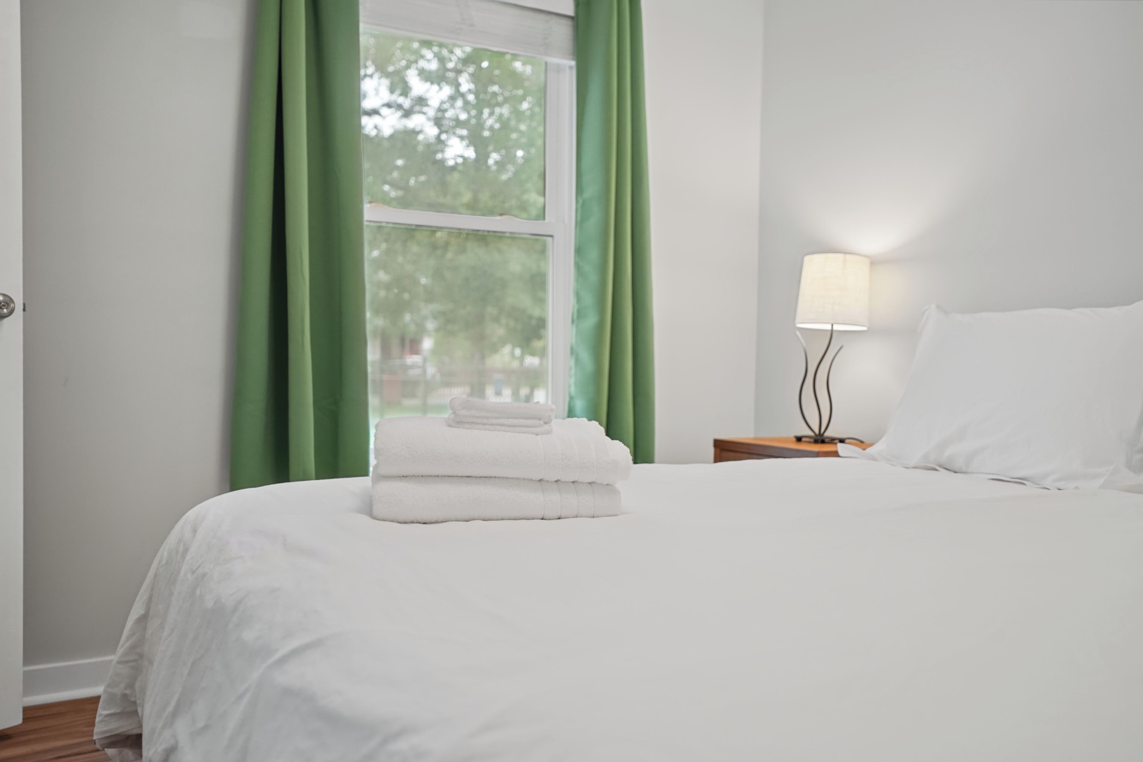 A neatly made bed with white linens and a stack of white towels on the foot of the bed. There is a bedside table with a lamp and a nightstand with a lamp. The room has a window with green curtains, and the walls are painted white.