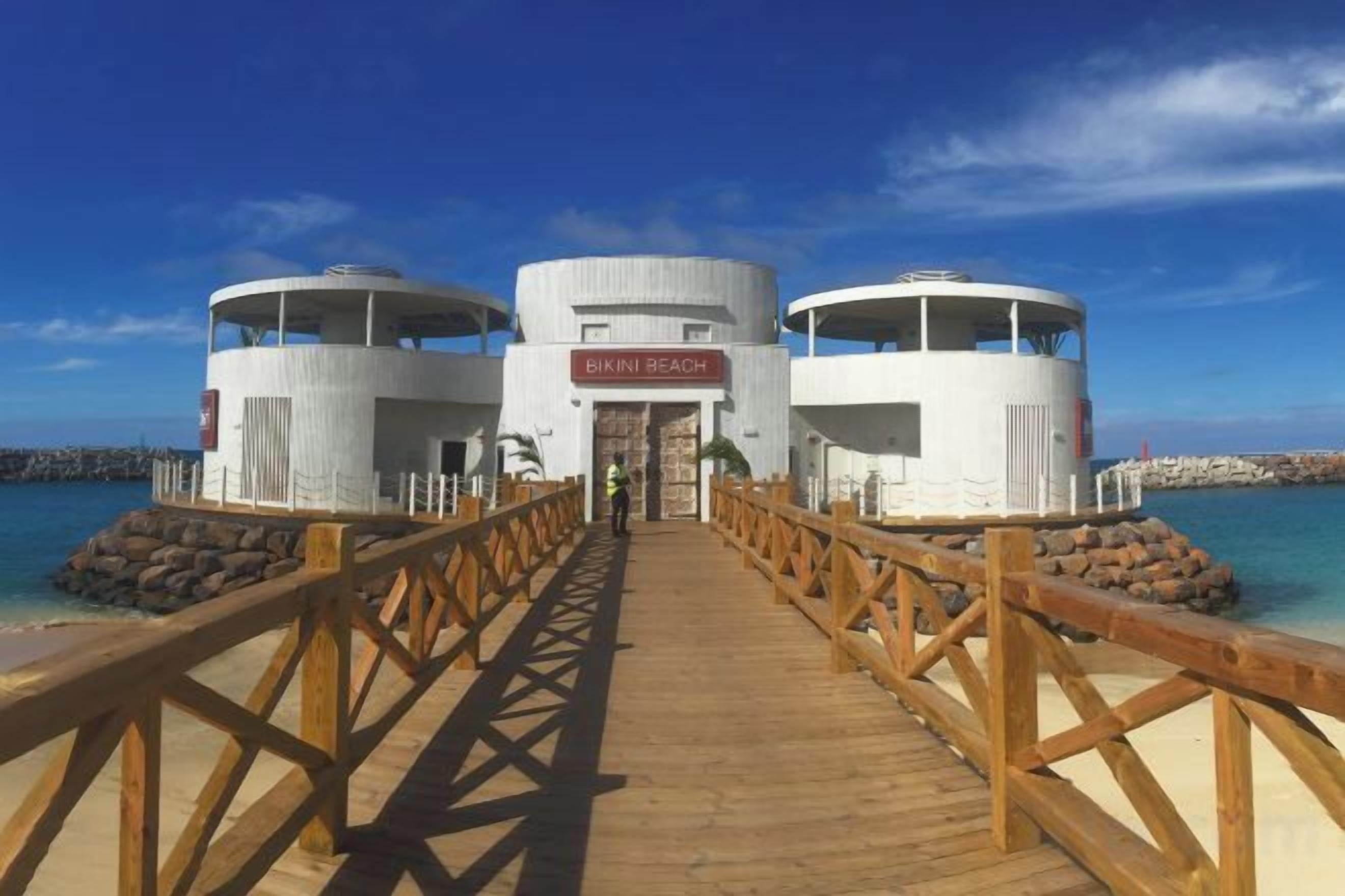Un muelle de madera que conduce a un edificio con el rótulo 'Bikini Beach', con una vista despejada del océano.