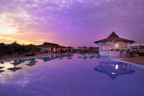 Una piscina con un pabellón y sillones de descanso, rodeada de sombrillas.