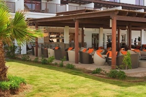 Un espacio de estar al aire libre con varios sofás y sillones, un porche cubierto y un edificio con balcón al fondo.