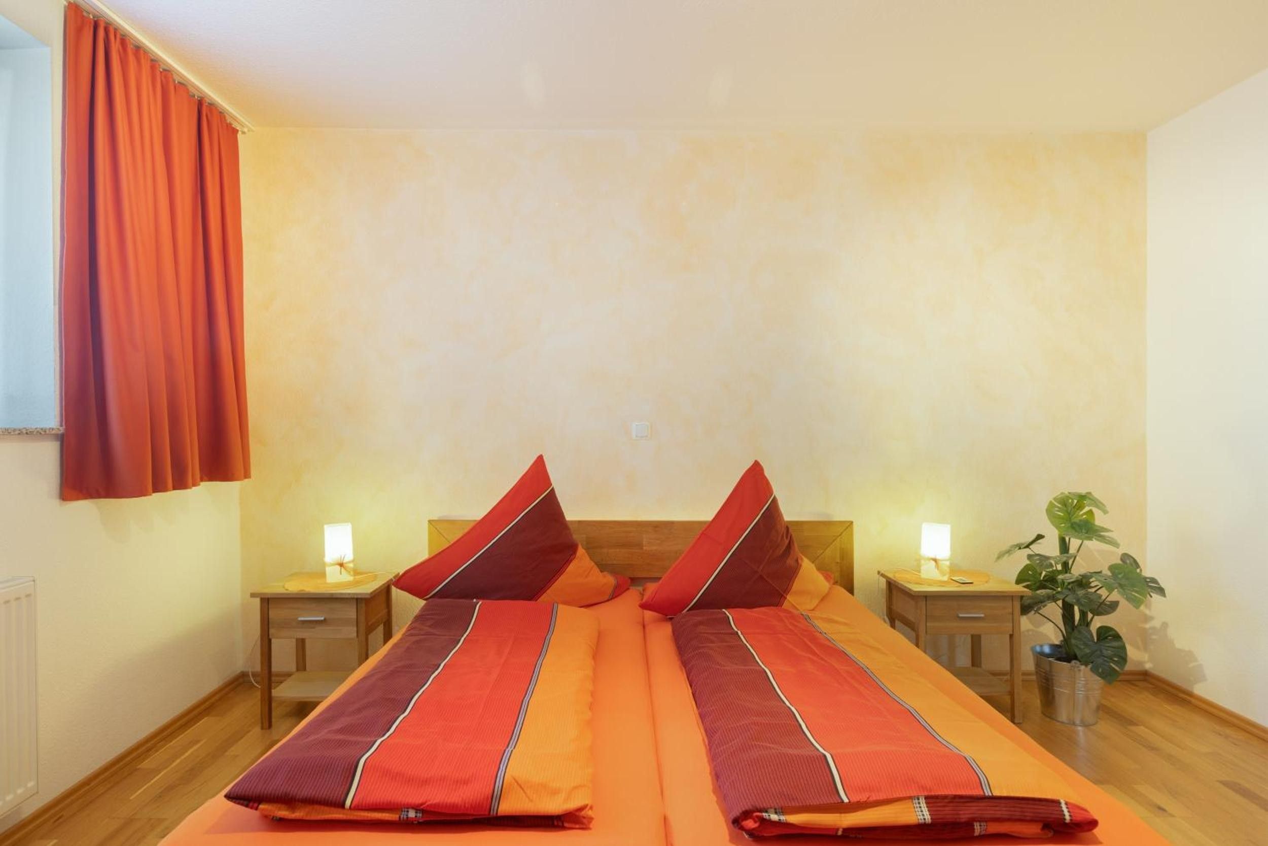 A double bed with matching pillows, two bedside tables with lamps, a potted plant, and a window with curtains.