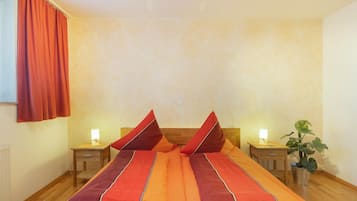 A double bed with matching pillows, two bedside tables with lamps, a potted plant, and a window with curtains.
