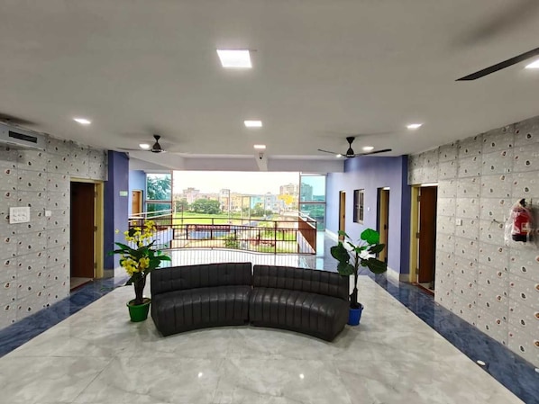 A hotel lobby with a curved black leather sofa, potted plants, a balcony with a railing, and a cityscape view.