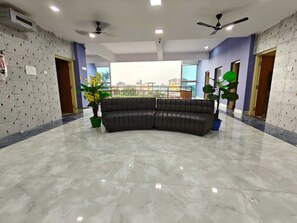 A spacious room with a curved black leather sofa, a large window offering a city view, and a ceiling fan. - Hotel O Atala Main Road (Balianta)