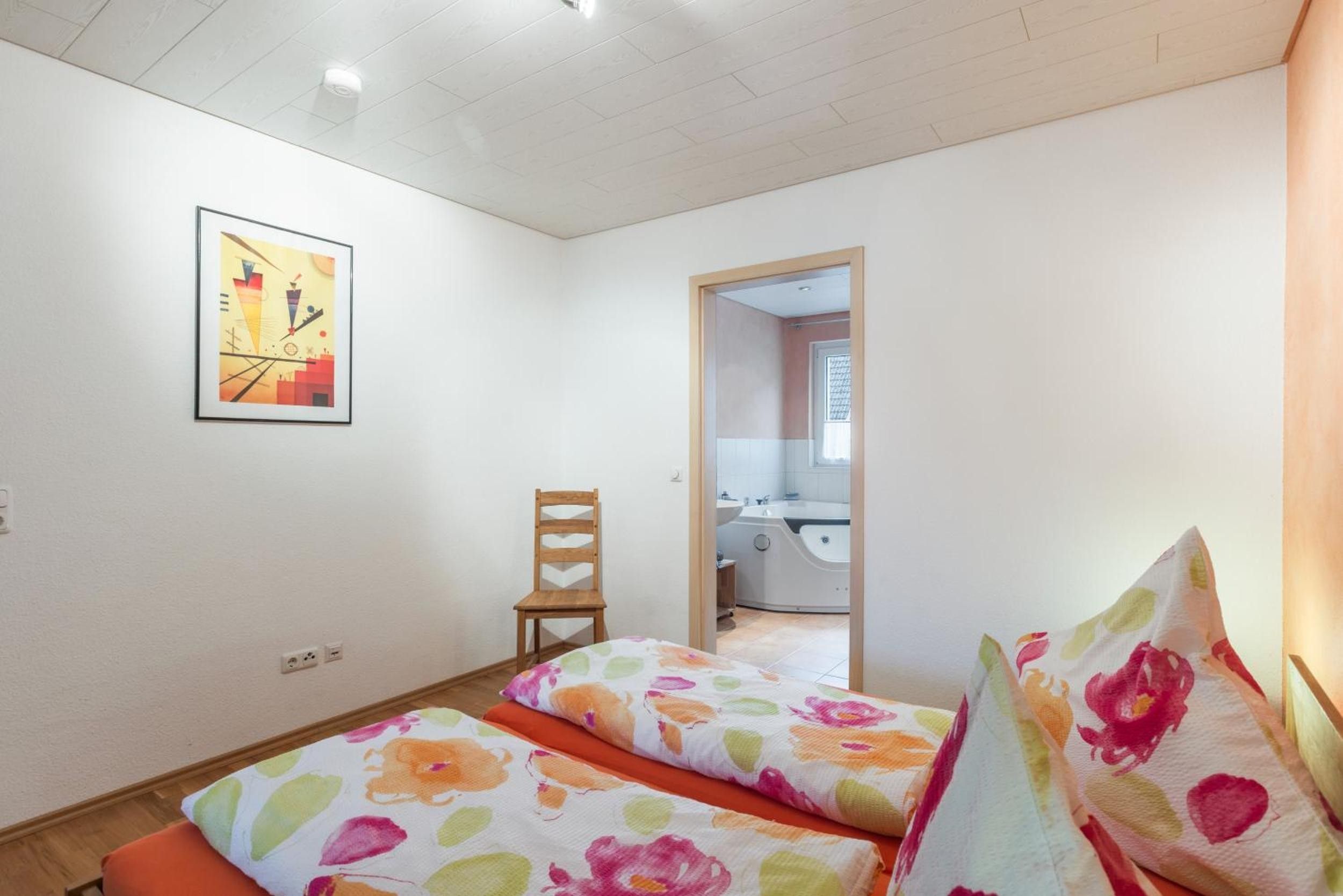 Una habitación con cama, una silla, un cuadro y un baño visible a través de una puerta abierta.
