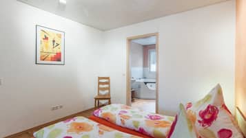 Una habitación con cama, una silla, un cuadro y un baño visible a través de una puerta abierta.