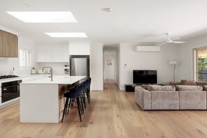 Et moderne køkken med en central ø, barstole, et spiseområde og en stue med sofa og tv.
