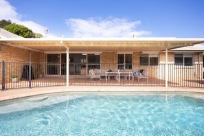 En swimmingpool med en overdækket terrasse, en murstensbygning og en klar blå himmel.