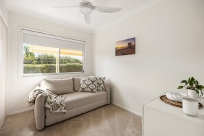 Una habitación con un sofá color beige, un ventilador de techo, un ventanal con vistas a zonas verdes, y una mesita con una planta y una vela.