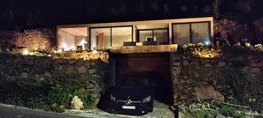 A modern house with a stone wall, a car parked in the driveway, and a well-lit interior. - Breathtaking views, peace and quiet, nature, and, of course, great waves! (São Vicente)
