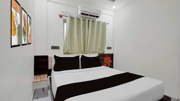A hotel room with a bed, a window with curtains, an air conditioning unit, and framed artwork on the wall.