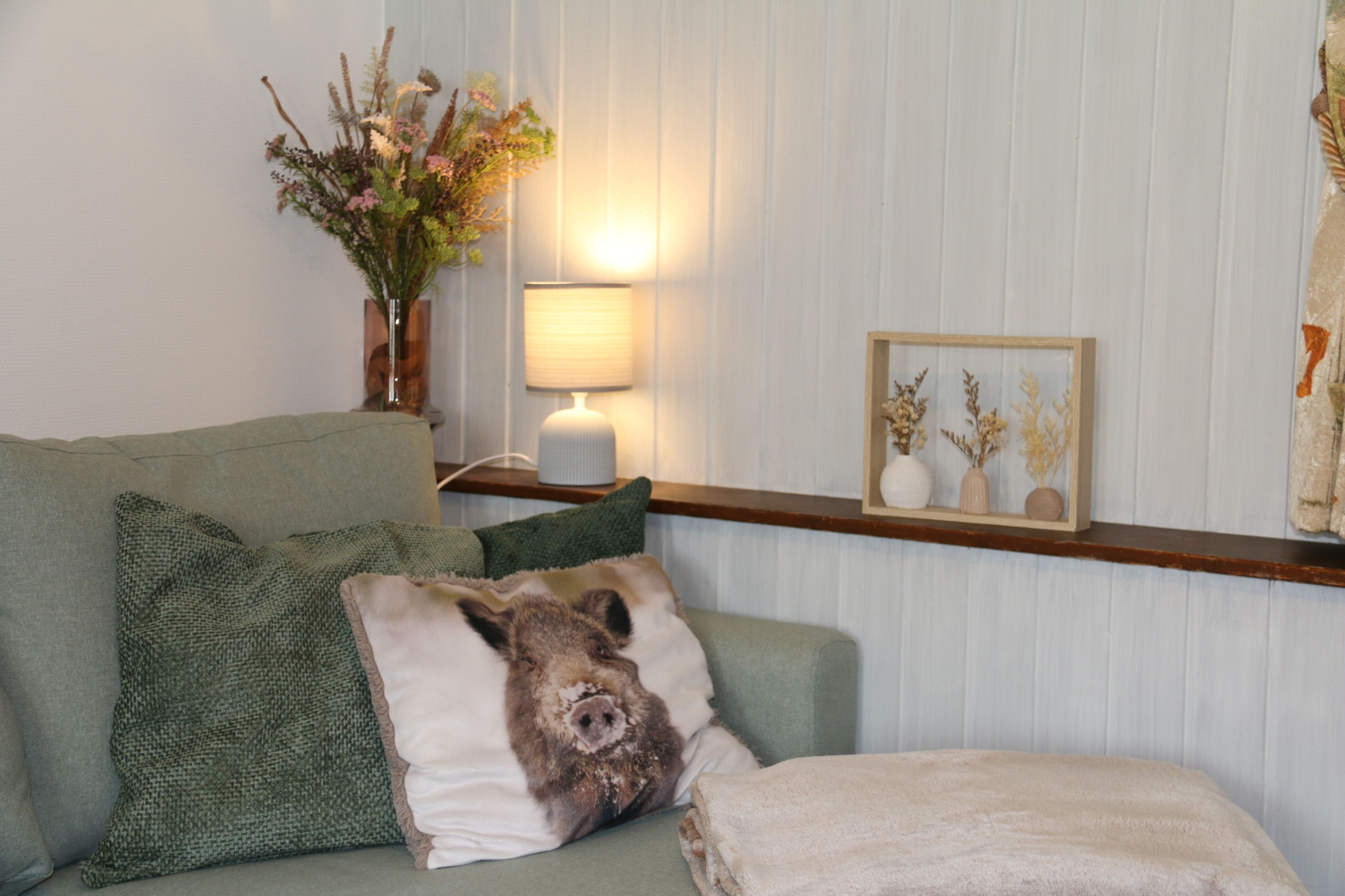 Ein Wohnzimmer mit einem grünen Sofa, einer Lampe, einem gerahmten Bild und einer Vase mit getrockneten Blumen.
