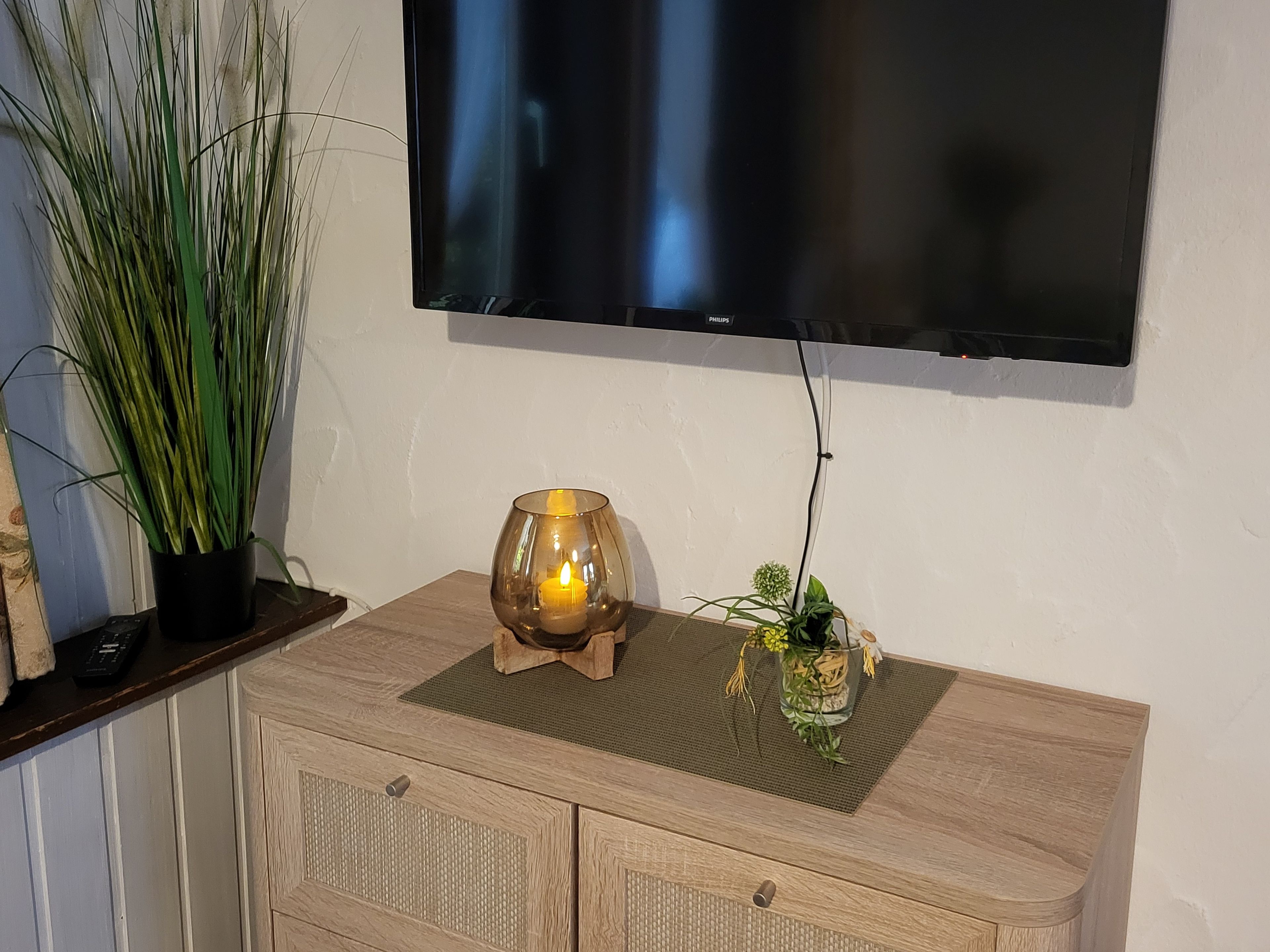 A television mounted on a wall, a wooden cabinet with drawers, a potted plant, and a candle holder with a lit candle.