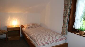 Une petite chambre avec un lit simple, une table de chevet, une lampe, une chaise et une fenêtre avec des rideaux.