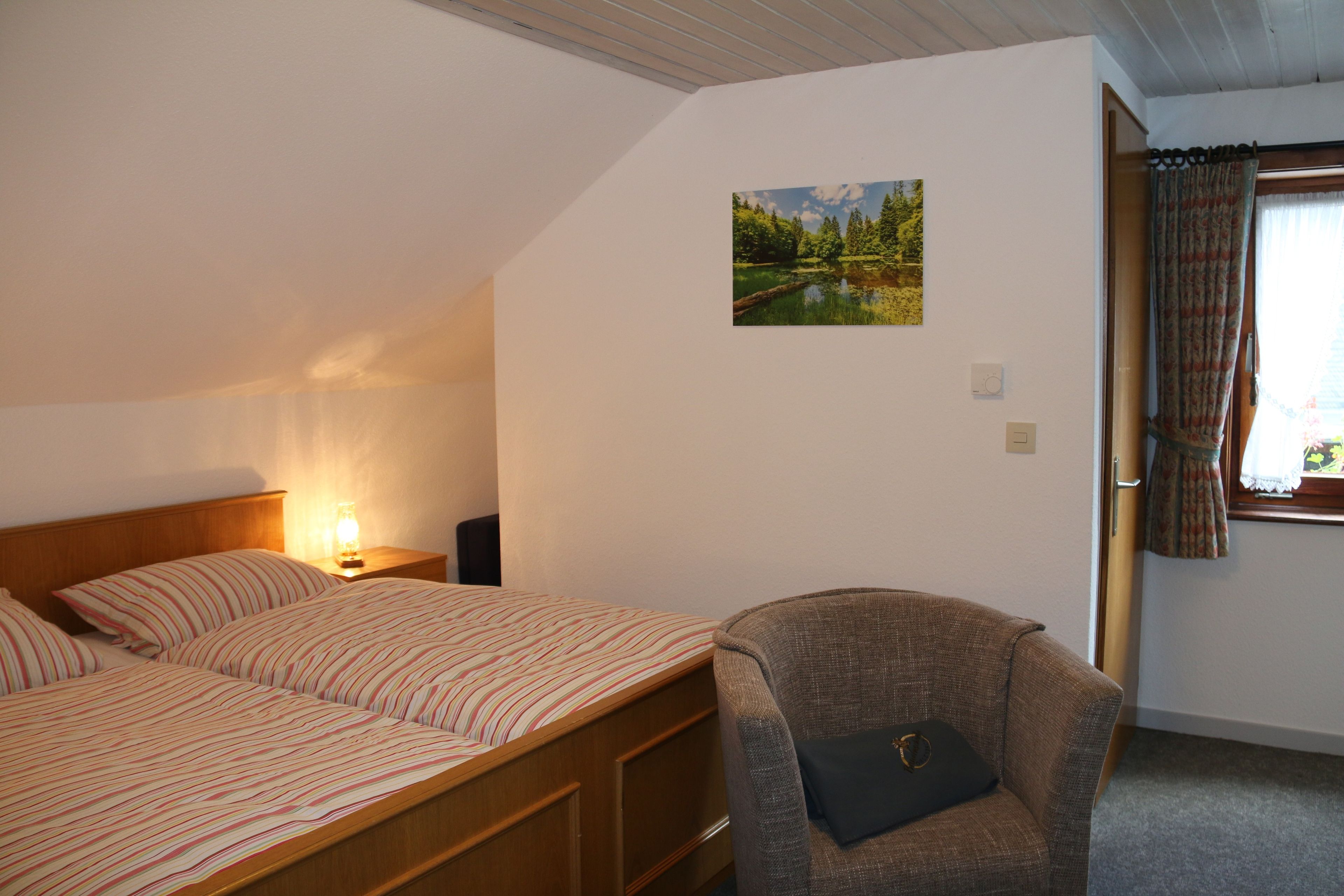 A bedroom with a bed, a chair, a nightstand with a lamp, a wall-mounted thermostat, and a window with curtains.