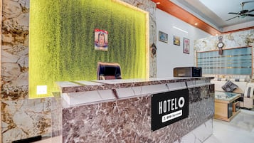 A hotel reception desk with a marble counter, a green wall with a poster, and a seating area with a sofa and a TV.