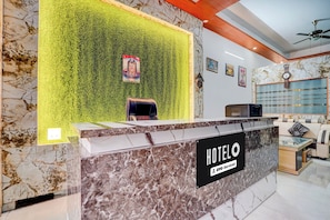 Mostrador de recepción de hotel con encimera de mármol, pared verde con un cartel y una zona de estar con sofá y televisor.