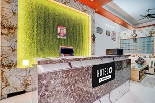 A hotel reception desk with a marble counter, a green wall with a poster, and a seating area with a sofa and a TV.