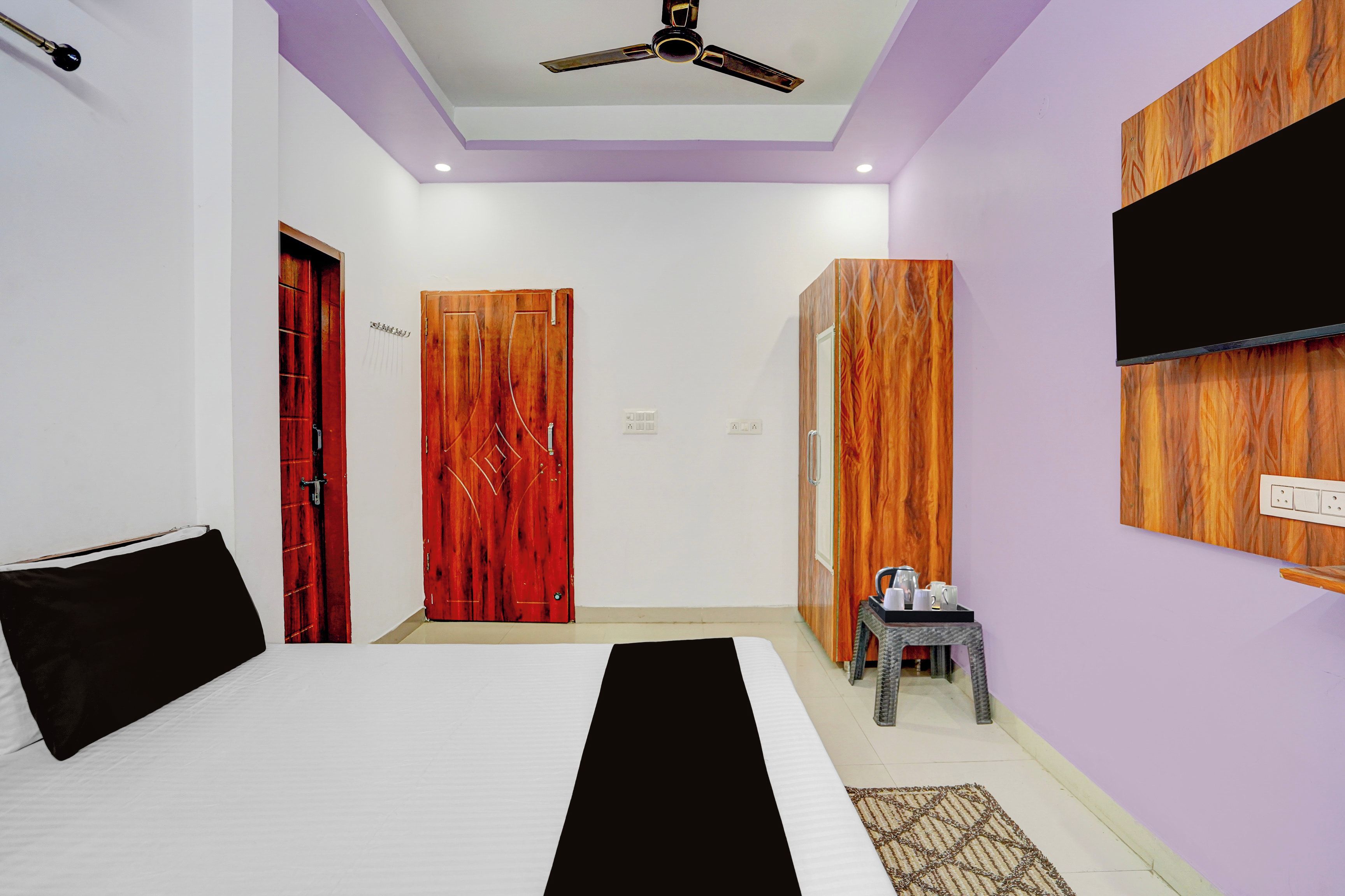 A hotel room with a bed, a ceiling fan, a TV mounted on the wall, and a wooden wardrobe.