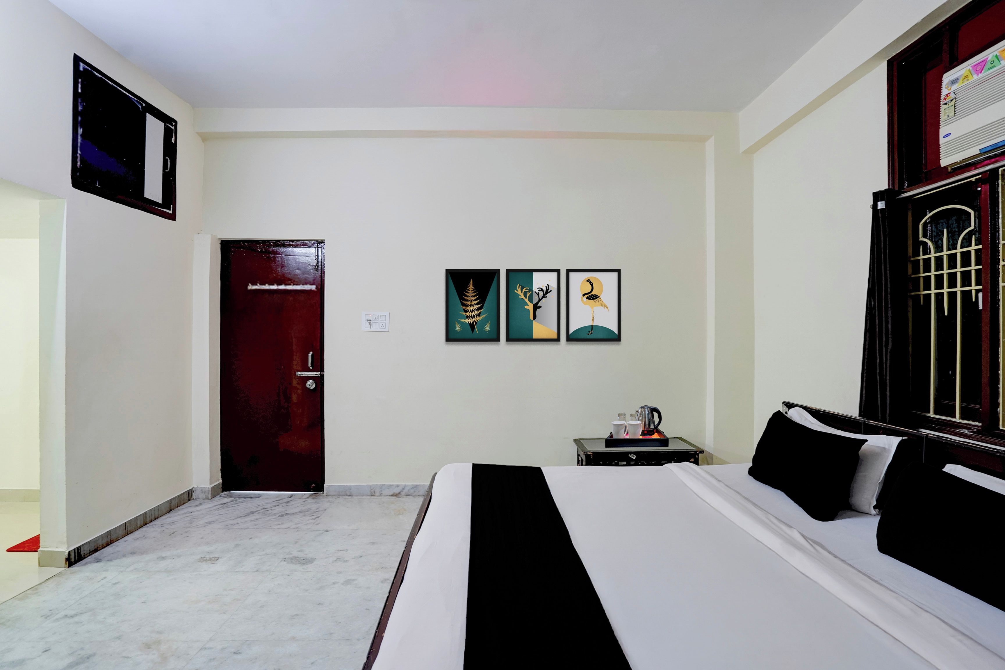 A hotel room with a bed, a desk with a coffee maker, and three framed artworks on the wall.