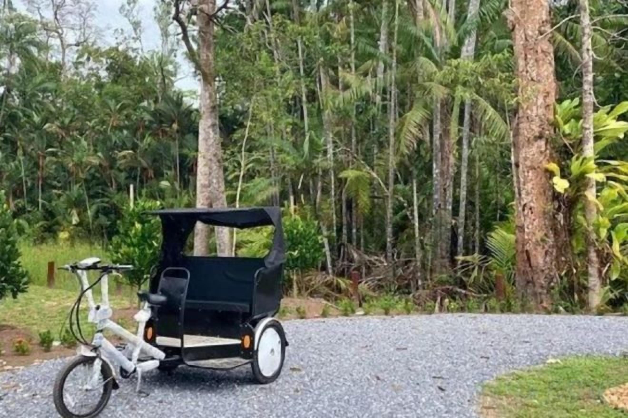 A white bicycle with a black trailer parked on a gravel path in a forested area.