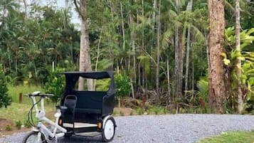 A white bicycle with a black trailer parked on a gravel path in a forested area.
