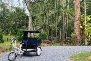 A white bicycle with a black trailer parked on a gravel path in a forested area.