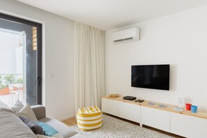 A modern living room with a sofa, a flat-screen TV mounted on the wall, a small wooden side table, and a window with curtains. - Apartment 'Light Filled' with Terrace (Porto)