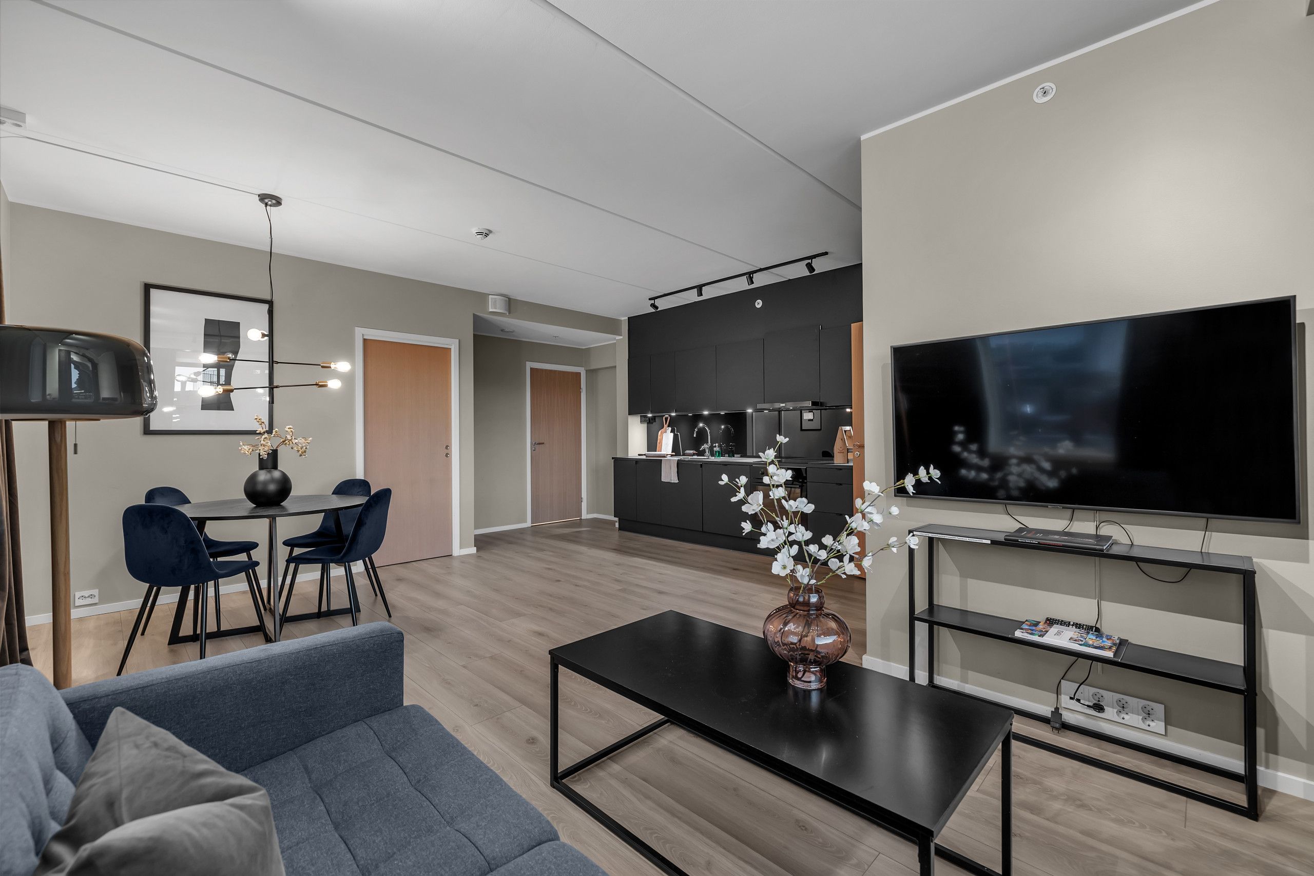 A modern living room with a sofa, coffee table, and a TV. There is a dining area with chairs and a table, and a kitchen area with cabinets and a refrigerator.