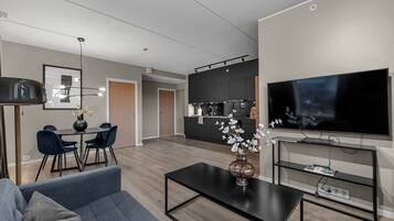 A modern living room with a sofa, coffee table, and a TV. There is a dining area with chairs and a table, and a kitchen area with cabinets and a refrigerator.