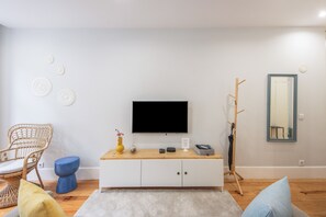 A modern living room with a white cabinet, a flat-screen TV, a wicker chair, a blue stool, and a wooden coat rack. - Apartment 'Cosy and Charming Almada Prime' (Porto)