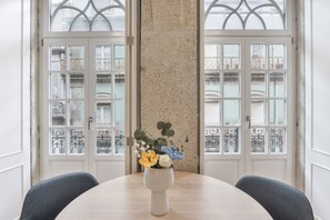 A dining table with a vase of flowers, two chairs, and a view through a window with a decorative iron railing. - Apartment 'Cosy and Charming Almada Prime' (Porto)