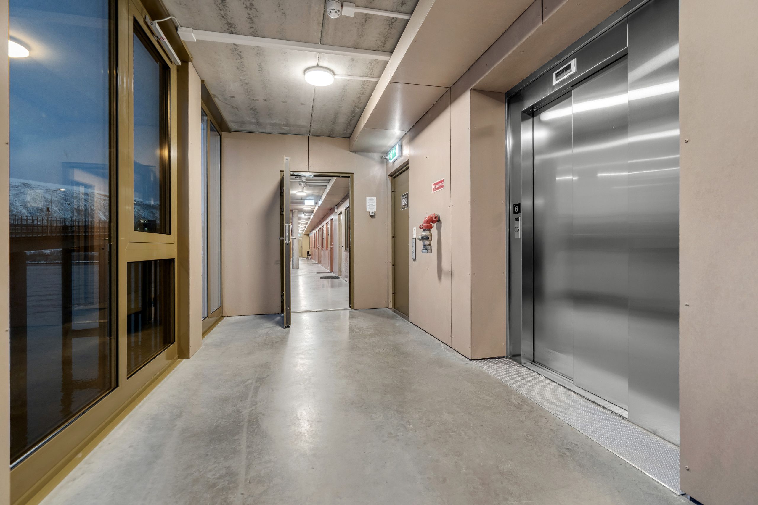 Un pasillo con piso de concreto, una puerta de vidrio a la izquierda y un ascensor a la derecha.