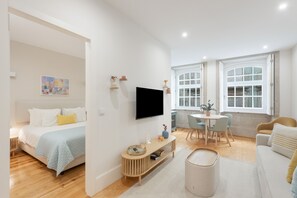A modern, well-lit room with a bed, a TV, a dining area with a table and chairs, and a window with a view. - Apartment 'Enchanting Almada Prime' (Porto)