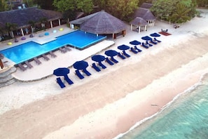 Vue aérienne d’un complexe hôtelier de bord de mer, avec une piscine, des chaises longues et des parasols.