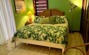 A bed with a leaf-patterned bedspread, a bedside lamp, a framed picture on the wall, and a window with blinds. - Blue Seas 11E Lavender Hill: Ocean Views, Pool & Walk-to-Town Location (St. John)