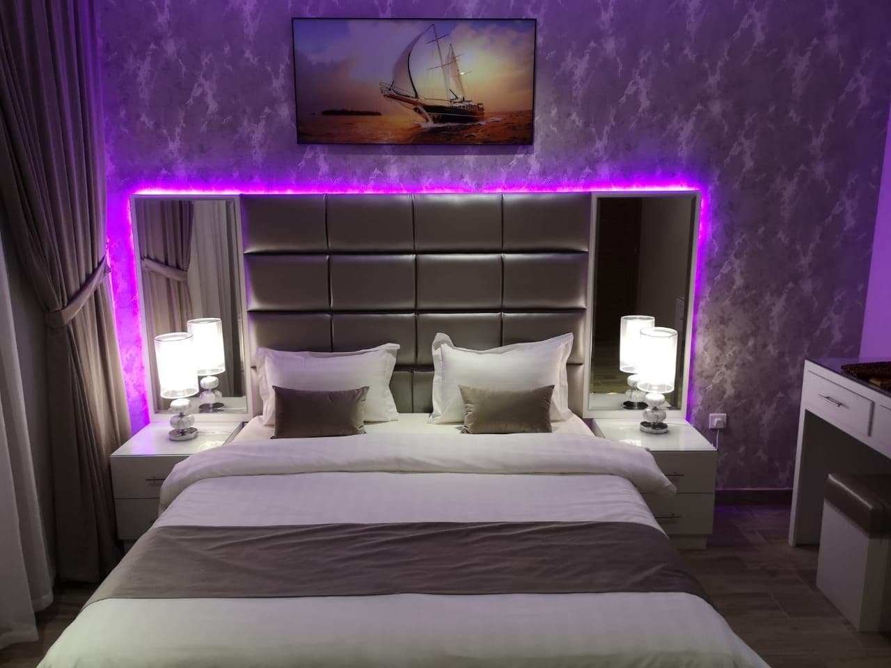 Una habitación de hotel con una cama grande, dos mesitas de noche con lámparas, un cabecero y un cuadro sobre la cama.